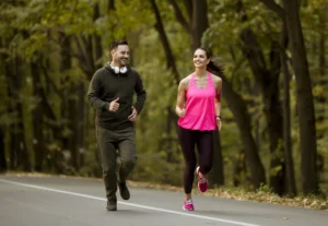 Happy young man and woman jogging together in nature on a tree-lined road, representing one of the "7 Things to Do Daily to Improve Your Life"—regular physical exercise. The uplifting scene highlights healthy habits, movement, outdoor activity, and emotional well-being.