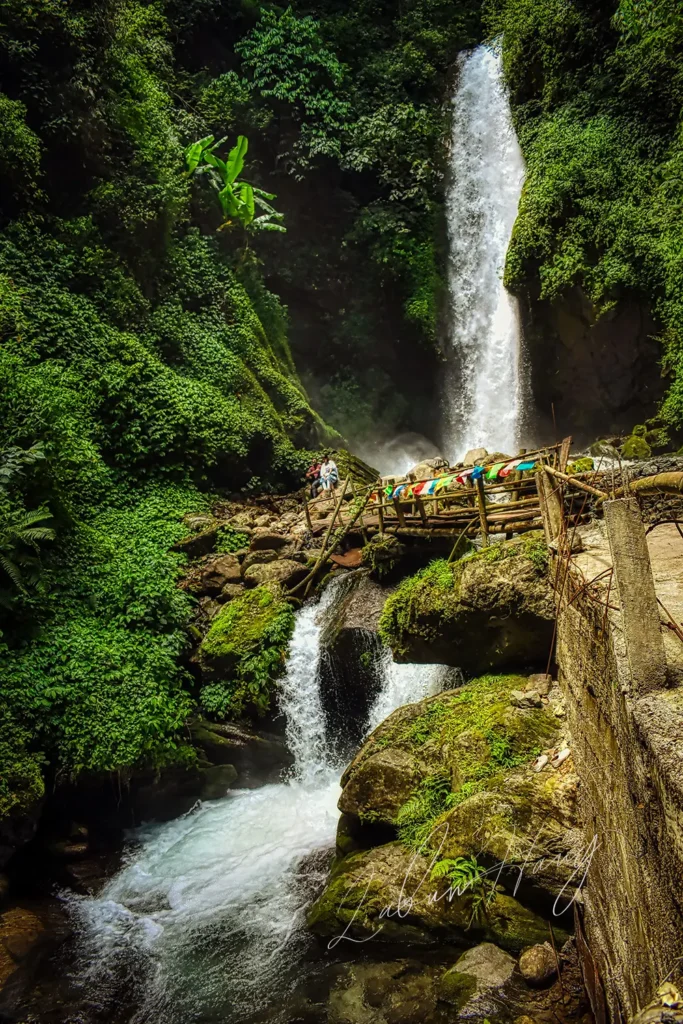 Kanchenjunga Waterfalls in West Sikkim showing cascading white water through lush green forest with wooden viewing bridge and colorful prayer flags