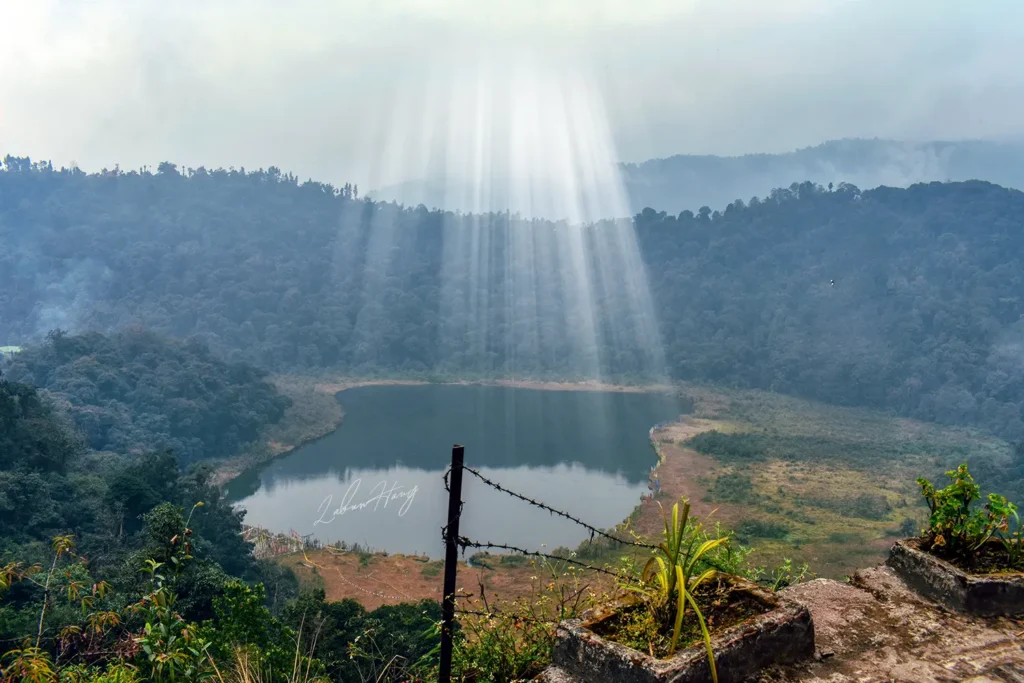 Khecheopalri Lake in West Sikkim with dramatic sunbeams piercing through misty clouds over the sacred Buddhist pilgrimage site surrounded by dense forest hills
